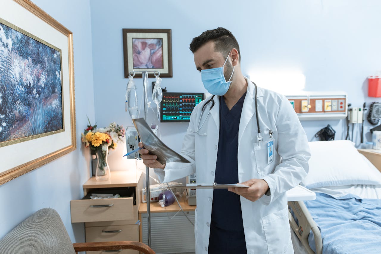 about-us A male doctor wearing a face mask reviews an X-ray in a hospital room, focusing on patient diagnosis.